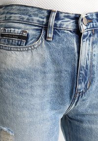 Los jeans de mezclilla azul claro presentan una textura desgastada con bordes deshilachados, un cierre de botón y una pequeña etiqueta de marca en el bolsillo delantero.