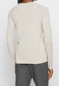 Ljusbeige långärmad topp med texturerad tyg, figursydd design och raglanärmar, kombinerad med mörkgrå byxor, som visar en avslappnad outfit.