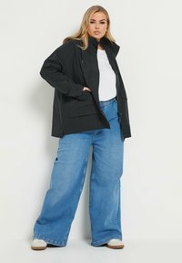 Svart vattentät jacka med dragkedja fram, justerbar huva och sidofickor, kombinerad med lösa blå denimjeans och vit topp.