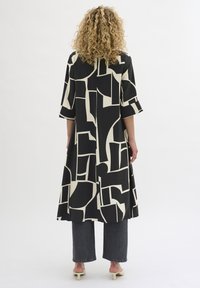 Abito geometrico in bianco e nero con maniche a tre quarti, forma fluida e lunghezza a metà polpaccio. Indossato con jeans scuri e scarpe con tacco.