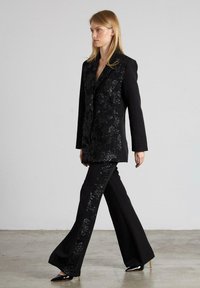 Blazer negro con lentejuelas y un corte elegante, combinado con pantalones negros acampanados que tienen un diseño de lentejuelas a juego a lo largo de los costados. Textura de tela suave.