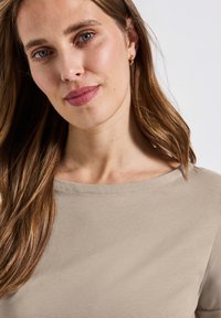 Béžový top s krátkým rukávem, širokým výstřihem a hladkou texturou, který nosí modelka s dlouhými vlasy a minimem doplňků.