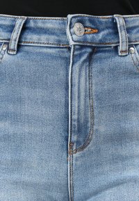Svijetloplave traperice s kopčanjem na dugme i kontrastnim narančastim šavovima. Ima pet džepova i ravni kroj.