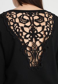 Top nero con dettagli in pizzo intricati sulla schiena, con un'apertura triangolare e motivi curvi. Texture del tessuto morbida.