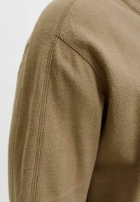 Acercamiento de una persona que lleva una camiseta de manga larga de color marrón claro con textura, mostrando detalles de las costuras en el hombro y la parte superior del brazo.