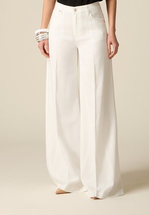 Donna che indossa pantaloni bianchi a vita alta e gamba larga con tasche frontali e scarpe beige con punta appuntita, accessoriata con numerosi braccialetti argentati e bianchi.