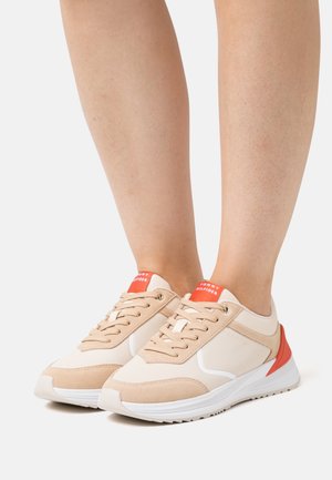 Beine in beige-weißen Tommy Hilfiger Sneakern mit orangefarbenen Akzenten und Schnürsenkeln, stehend auf einem schlichten hellen Hintergrund.