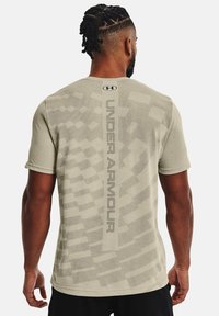 Man med flätat hår bär en ljusbeige Under Armour T-shirt med mönstrad baksida och vertikal logotyp, vänd bort, med avslappnade händer vid sidorna.