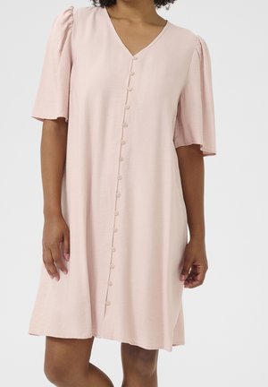 Kvinde iført en lyserød kortærmet kjole med V-hals og række af knapper foran, stående mod en ensfarvet hvid baggrund.