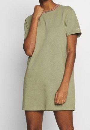 Kvinde iført kortærmet, olivengrøn, afslappet kjole med Tommy Hilfiger-logo på ærmet og mærket rund hals står mod en ensfarvet baggrund.