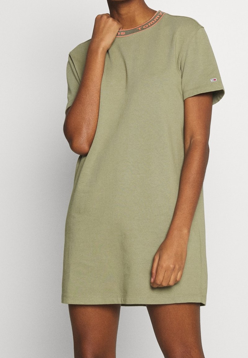Femme portant une robe décontractée courte à manches courtes de couleur vert olive avec le logo Tommy Hilfiger sur la manche et un col rond marqué, se tenant devant un fond uni.