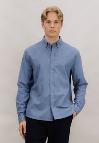 Lys blå skjorte med knapper laget av vevd stoff, med klassisk krage, lange ermer og rett kant med fem knapper foran.