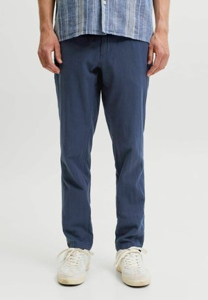 Mann trägt marineblaue Hose, weiße Sneaker und ein blau-weiß gestreiftes Hemd mit Knöpfen und steht vor einem einfarbigen Hintergrund.