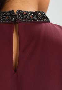 Encolure en tissu bourgogne avec des perles complexes accentuées de noir et de cuivre. Une petite ouverture à l'arrière ajoute un détail de design.
