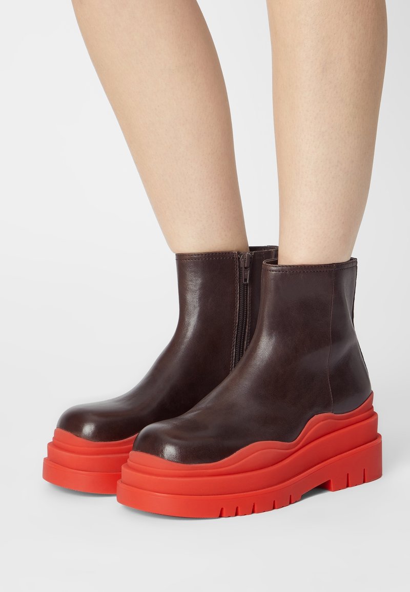 Bottes basses en cuir marron avec semelles plateforme épaisses rouges vives, portées sur des jambes nues devant un fond uni.