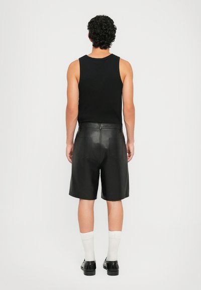 Man with curly hair wearing a black sleeveless top, black leather shorts, white socks, and black shoes, standing facing away on white background.