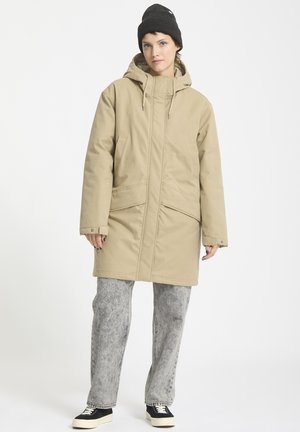 Beige gewatteerde jas met een capuchon, voorzien van voorzakken en een ritssluiting. Gepaard met grijze jeans en zwarte sneakers.