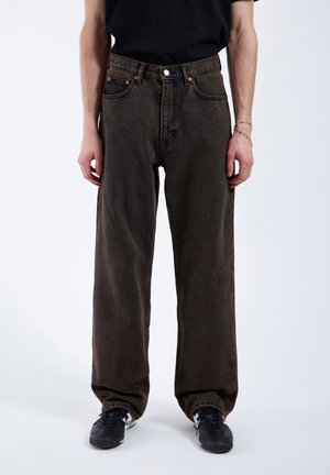 Wijd-uitlopende, donkerbruine denim jeans met een hoge taille, vijf zakken en contrasterende gouden stiksels. Draagt met zwarte sneakers.