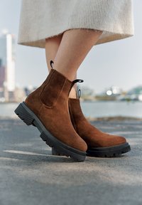 Botas de ante marrón hasta el tobillo con una suela de goma negra gruesa, que cuentan con una pestaña para tirar y paneles laterales elásticos para facilitar el uso.