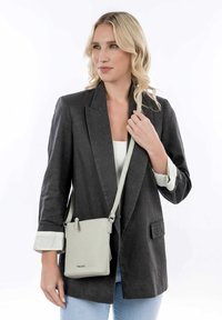 Borsa a tracolla in pelle grigio chiaro con tracolla regolabile, caratterizzata da una superficie texturizzata e angoli arrotondati, indossata con un blazer scuro.