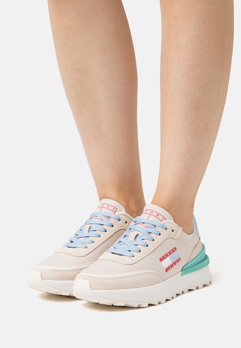 Personne portant des baskets beige clair avec des lacets bleu clair, un logo rouge et blanc, et un détail talon bleu sarcelle sur fond blanc.
