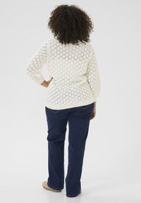 Witte gebreide trui met diamantpatroon, gebreide details en driekwartmouwen, gedragen met donkerblauwe jeans. Achteraanzicht van de outfit.