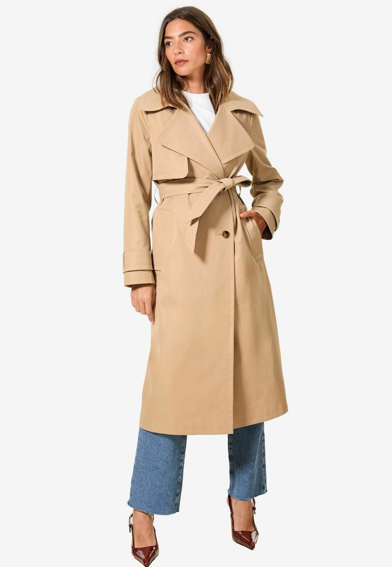 Abrigo trench beige con solapas anchas, cintura ceñida y bolsillos; presenta una textura suave y un diseño hasta la rodilla, acompañado de jeans azul cortos.