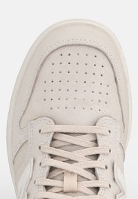 Sneaker beige avec bout perforé, lacets tressés et semelle blanche, vue de dessus sur un fond uni.