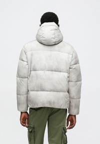 Gaisma pelēka pūkainā jaka ar kapuci, ar quiltētām daļām un gludu tekstūru. Saskaņota ar olīvzaļām militārā stila biksēm.