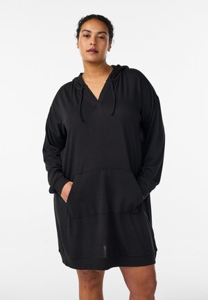 Žena v čiernych voľných šatách s kapucňou, dlhými rukávmi a predným kangaroo vreckom, stojaca na jednoduchom bielom pozadí.