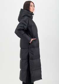 Manteau noir long en duvet avec capuche, présentant un design matelassé, des poches latérales et un ourlet fendu pour faciliter les mouvements. Texture lisse et brillante.