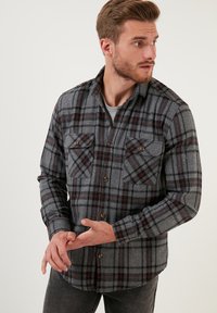Camicia in flanella a quadri grigi e rossi con due tasche sul petto, bottoni marroni e un orlo leggermente curvato. Maniche lunghe con polsini abbottonati.