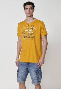 Geltona trumpomis rankovėmis marškinėliai su Henley apykakle, ant kurių yra grafiškas tekstas baltos ir tamsiai mėlynos spalvos. Dėvimi su mėlynais džinsiniais krovinių šortais.