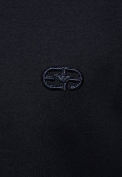 Μαύρο ύφασμα με ένα μικρό, σκούρο κεντητό λογότυπο που απεικονίζει έναν οριζόντιο οβάλ σχήμα και αφηρημένο γεωμετρικό σχέδιο στο εσωτερικό.