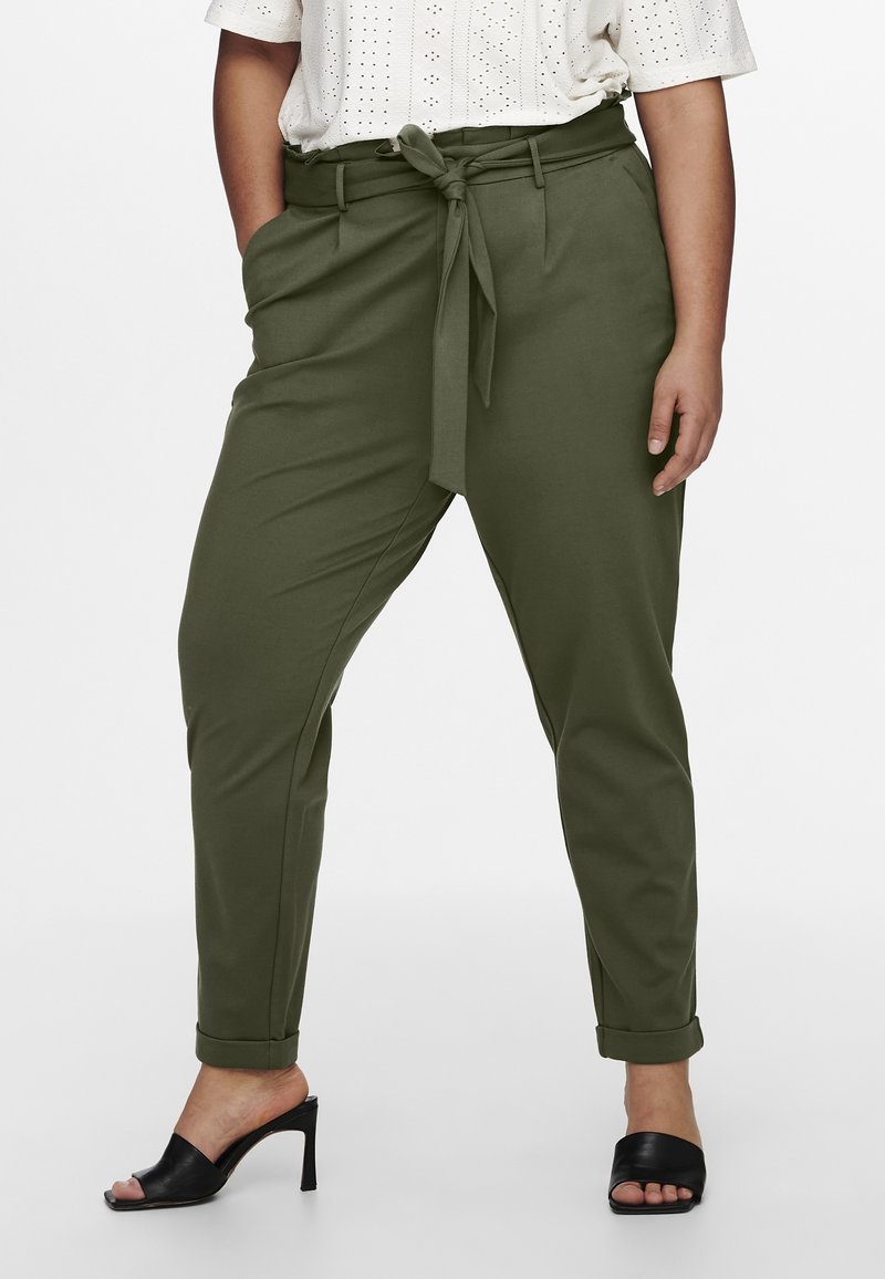 Femme portant un pantalon fuselé taille haute vert olive avec une ceinture nouée à la taille, associé à des mules noires à talons ouvertes à l'avant et un top blanc à œillets.