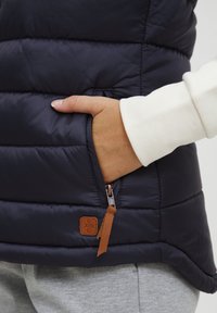 Tamno plavi puffa prsluk s kvadratnim šavovima, s bočnom džepom sa zatvaračem i bež kožnom oznakom. Glatka, sjajna tekstura.