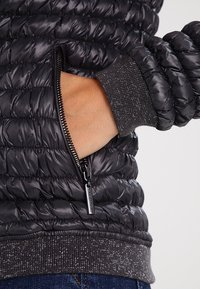 Zwarte pufferjack met een doorstikte textuur, een ritssluiting zijzak en een grijze geribbelde manchette met metallic deeltjes. Zacht, lichtgewicht materiaal.