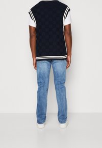 Veste en tricot bleu marine à motif damier, rayures blanches contrastantes aux épaules et à l'ourlet, portée sur un t-shirt blanc, associée à un jean bleu clair.
