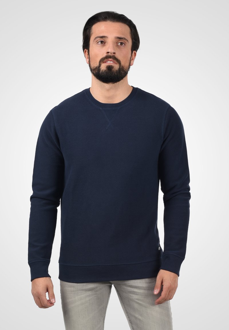 Mörkblå sweatshirt med långa ärmar, rund halsringning och strukturerad tyg. Har ribbade mudd och nederkant, med en liten logotypdetalj i botten.