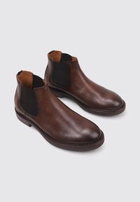 Bottines en cuir marron avec des panneaux latéraux élastiques, bouts ronds et accents texturés sur une surface lisse. Semelle plate et détails de couture subtils.
