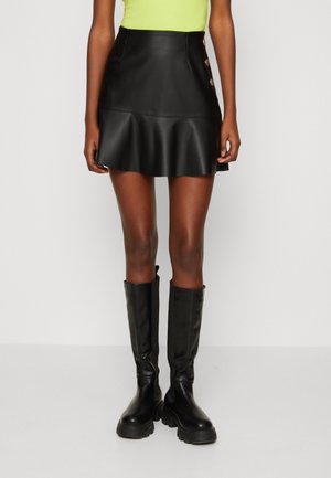 Mujer con minifalda de cuero negra con botones laterales, botas negras hasta la rodilla y una blusa amarillo neón, de pie contra un fondo blanco.