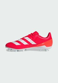 Botín de fútbol de material sintético en un brillante color rojo, con franjas blancas, punta redondeada y suela blanca y naranja con tacos moldeados.