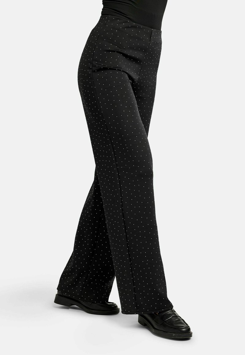 Mustad laia säärega püksid väikeste valgete täppidega, mida kannab mustade loaferitega inimene, kes seisab valge tausta ees.