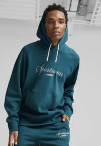 Felpa con cappuccio color teal, dotata di cordini regolabili, tasca anteriore e logo ricamato "Sportswear by PUMA". Tessuto morbido e strutturato.