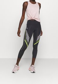 Svarta sportleggings med vita och neongröna diagonala ränder, kombinerat med en rosa knuten linne, och ljusrosa löpskor.