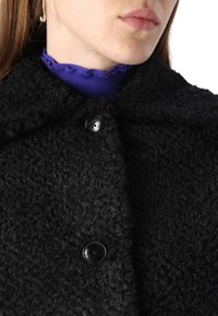 Outerwear nero con texture, dotato di colletto e dettagli con bottone lucido. Sotto la giacca è visibile una maglietta a coste viola.