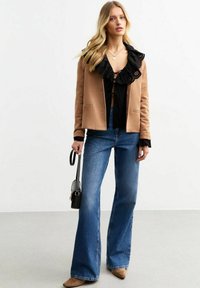 Brun kavaj över en svart volangblus, kombinerat med vida blå jeans och beige skor. Accessoarer inkluderar en liten svart handväska.
