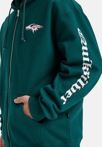Tyrkysová mikina na zips s plamenným logom na hrudi a bielym textom "Quiksilver" na rukáve, ruka osoby čiastočne v prednom vrecku.