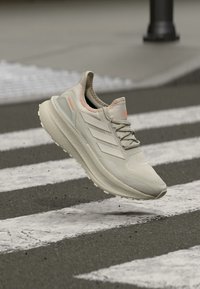 Beiger Sportschuh mit einer strukturierten Oberseite, auffälligem Drei-Streifen-Branding und einer gepolsterten Sohle. Verfügt über einen pinkfarbenen Akzent rund um den Kragen.