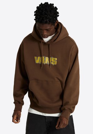 Persona con sudadera marrón con capucha y texto amarillo "VANS" envuelta en alambre de púas, mirando hacia abajo con las manos relajadas a los lados.